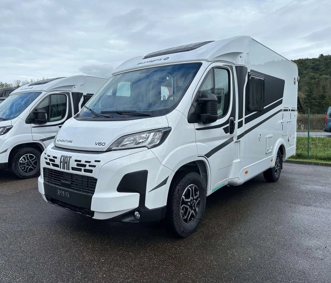 Camping-car Fiat V60 blanc stationné sur un parking, ciel nuageux en arrière-plan.