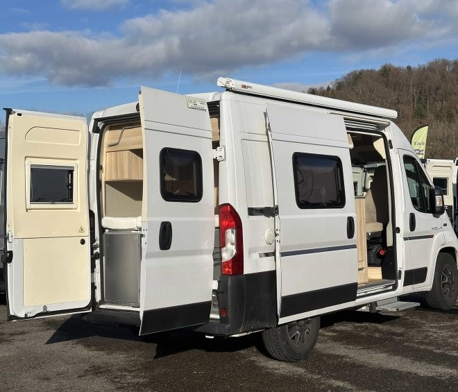Fourgon aménagé blanc avec portes ouvertes sur un parking, ciel bleu en arrière-plan