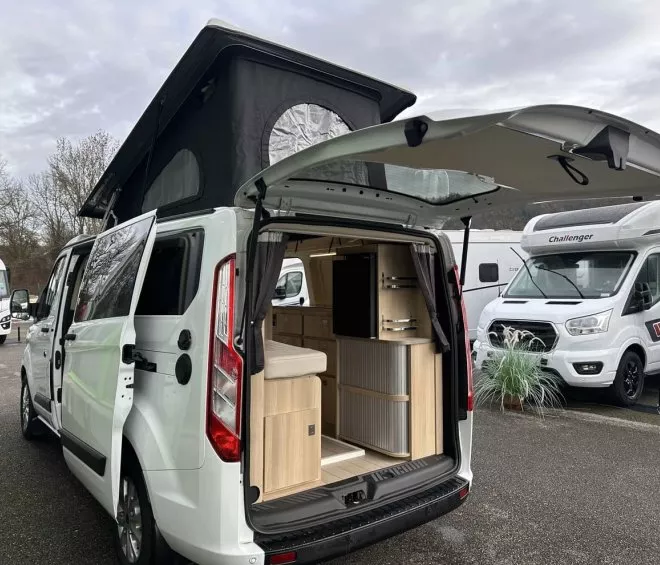 Van aménagé avec toit relevable et intérieur visible, stationné sur un parking avec d'autres camping-cars.