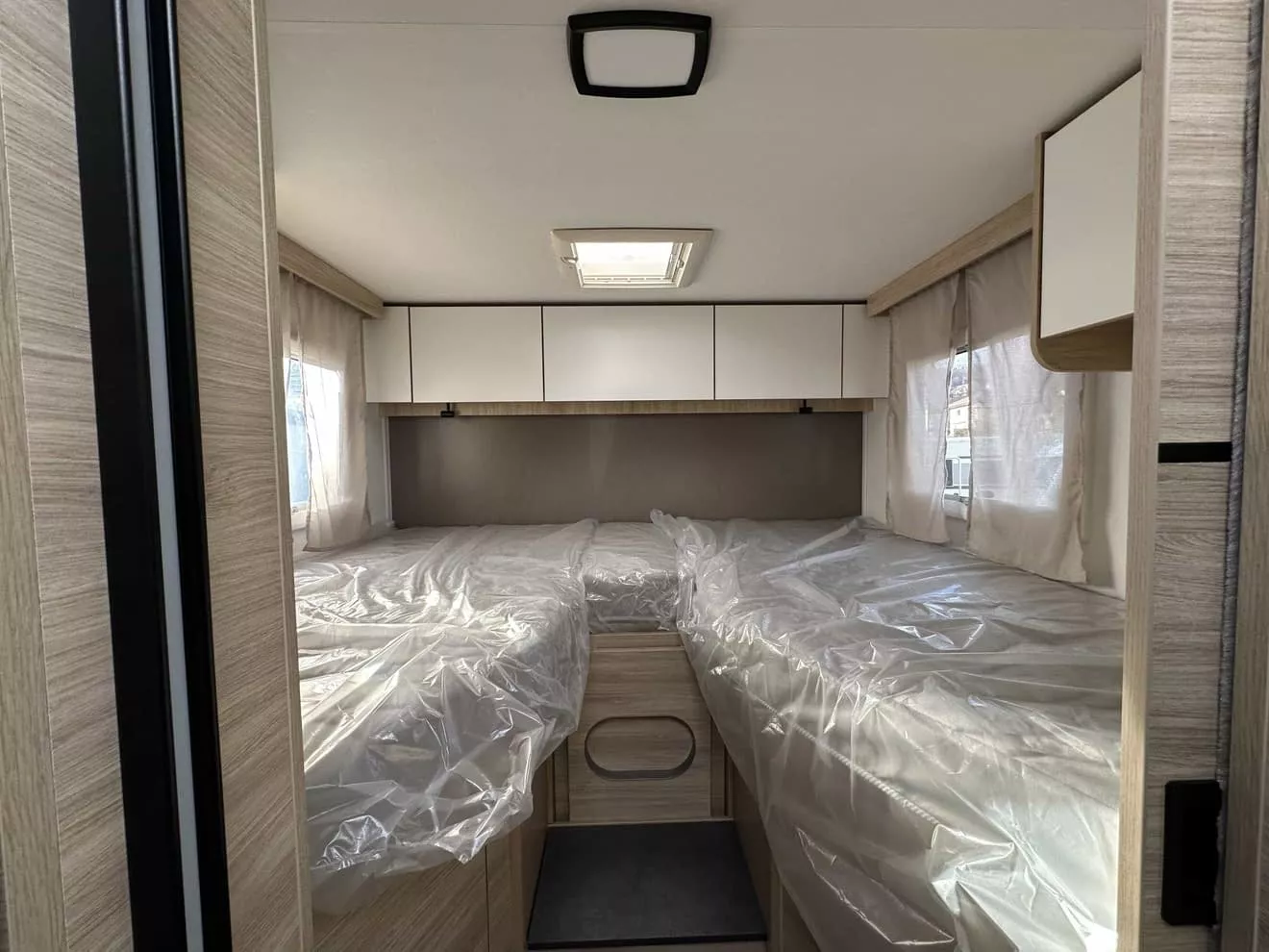 Intérieur d'une caravane avec lits jumeaux sous plastique, fenêtres latérales et placards.