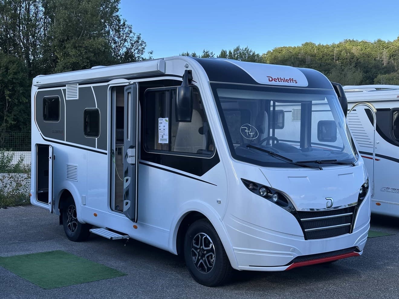 Camping-car Dethleffs blanc stationné sur un parking, porte ouverte, ciel bleu en arrière-plan.