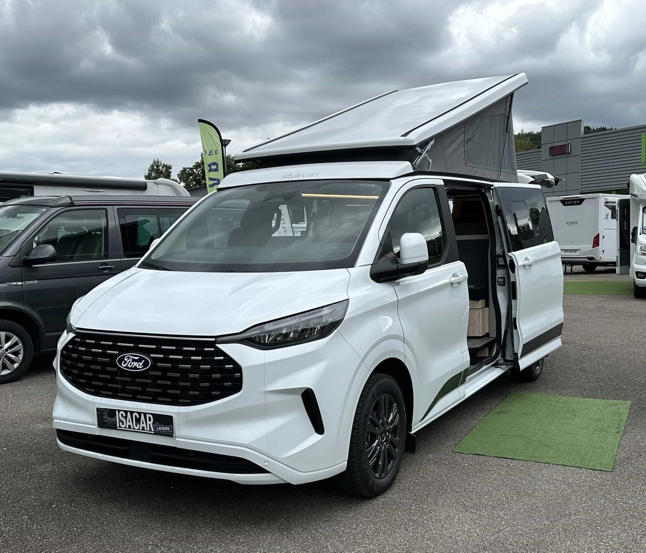 Van aménagé Ford blanc avec toit relevable sur un parking de concessionnaire.