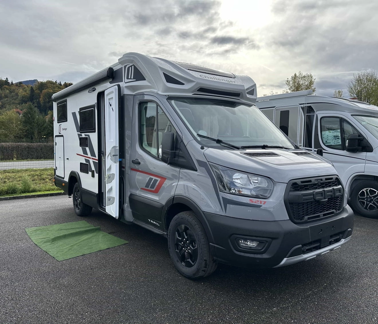 Camping-car gris stationné sur un parking, porte ouverte, ciel nuageux en arrière-plan.