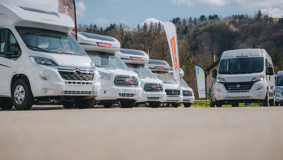 Alignement de camping-cars blancs sur un parking, bannières publicitaires en arrière-plan.